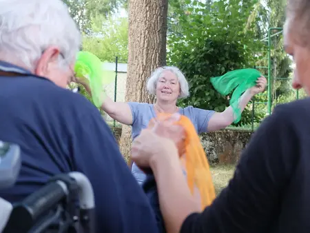 Dorothee Ewald steht strahlend vor Senior*innen und hält zwei grüne Tücher in die Höhe.